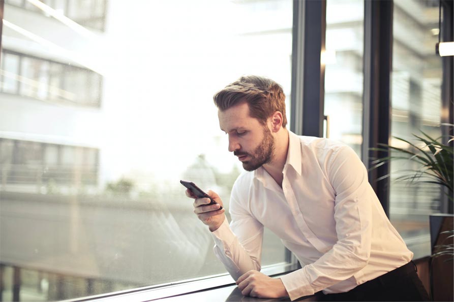 A photo of a man in a white dress shirt checking his phone, leaning by a window